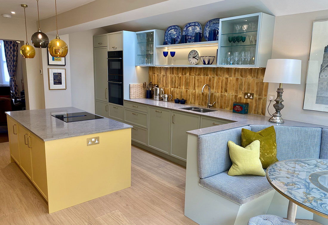 The bespoke designed kitchen in Wandsworth in sage green with contrasting yellow island, marble tops and glass pendant lamps.